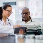 A senior man of African descent is indoors in a hospital room, looking at a tablet computer with his doctor. The doctor, a woman, is explaining his medication schedule to him.