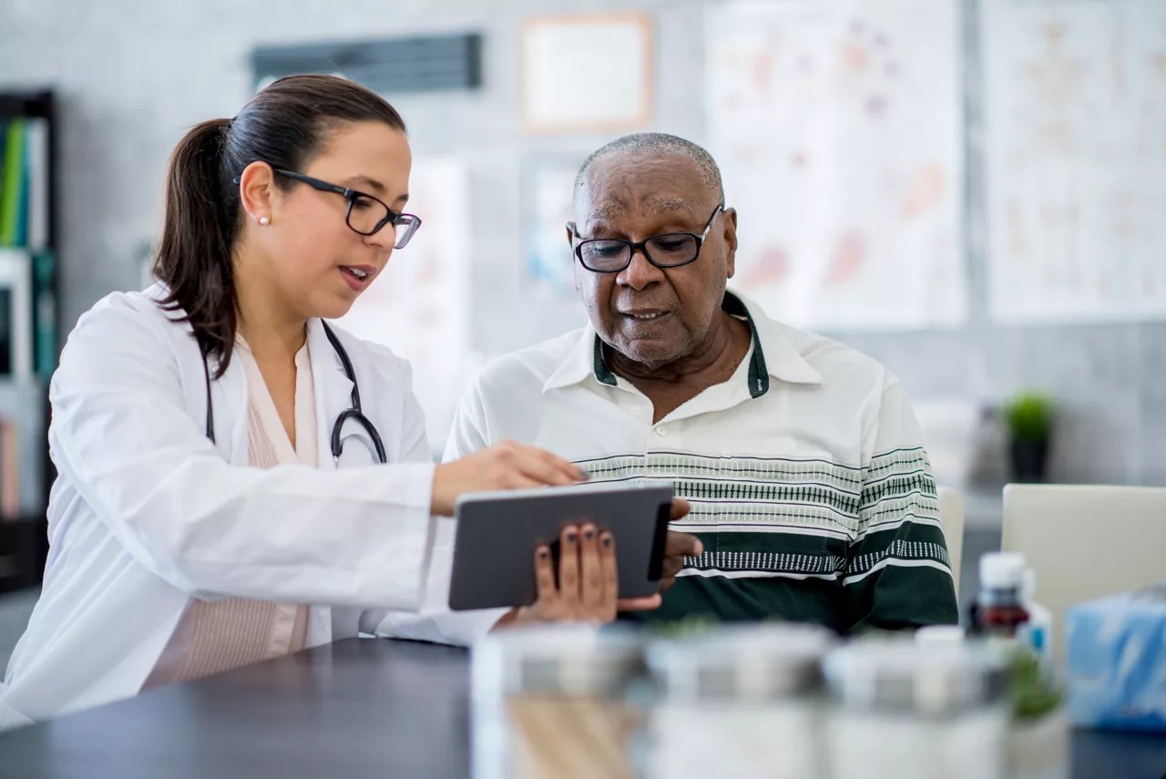 A senior man of African descent is indoors in a hospital room, looking at a tablet computer with his doctor. The doctor, a woman, is explaining his medication schedule to him.